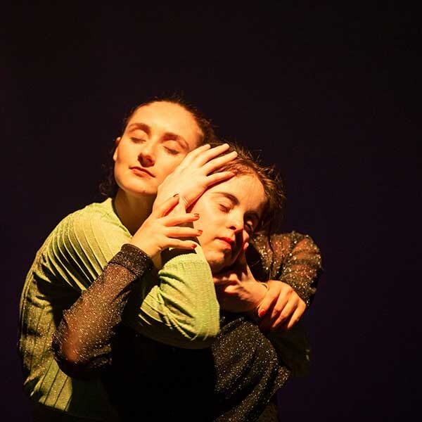 Two people stand against a pitch black background and in direct lights with their eyes closed, cradling each other's faces.