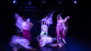 Michelle Ryan seated in her chair while her company of Restless dancers move around her. You can see the dancers moves are blurred but Michelle is not as she sits completely still.