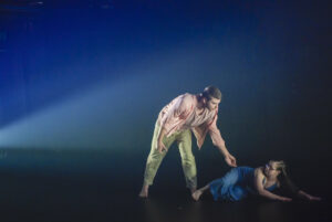 A man in a pink shirt reaching down to a woman lying on the ground in a blue dress.