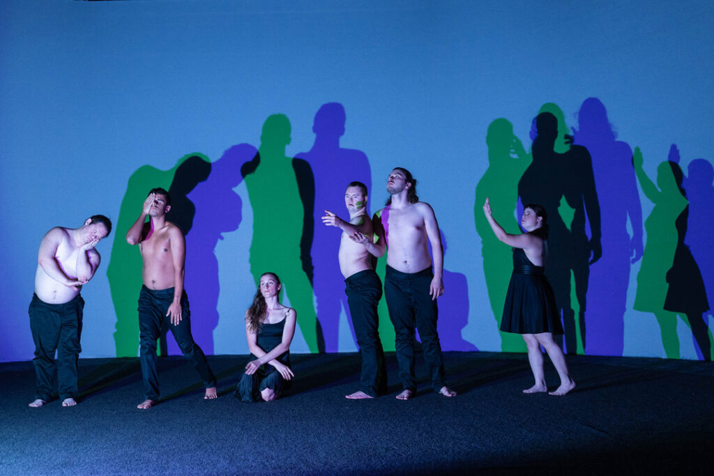 and ensemble of dancers all posing like statues with blue light reflecting different colours in their shadows behind them.