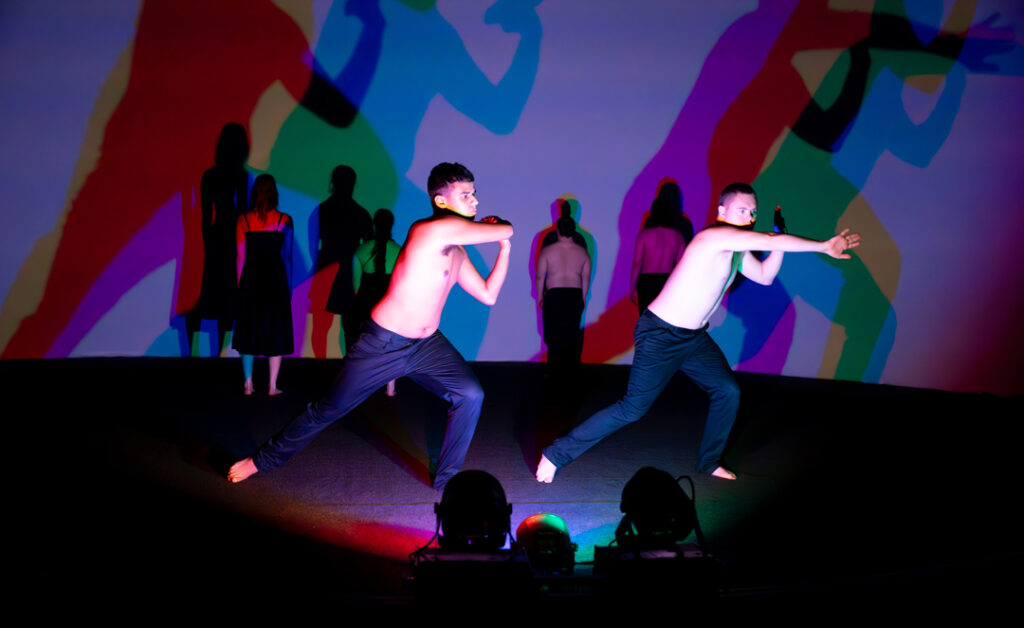 Two male dancers in a strong swirling dance move. Purple light refracting different colours behind them.