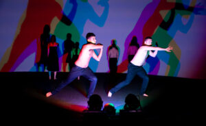 Two male dancers in a strong swirling dance move. Purple light refracting different colours behind them.
